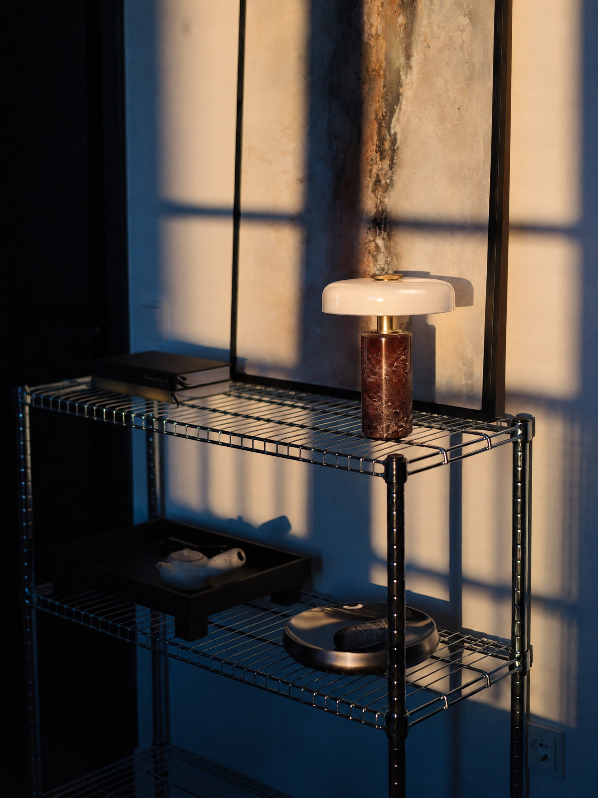 The Columba lamp unlit on a chrome wire shelf in evening sun, window shadows falling across a weathered artwork behind it.