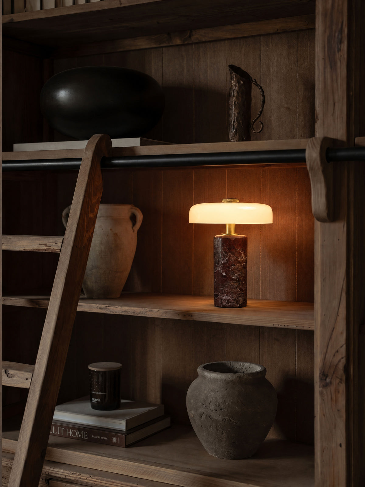The Columba lamp lit on a reclaimed-wood library shelf behind a wooden ladder, beside a terracotta amphora.