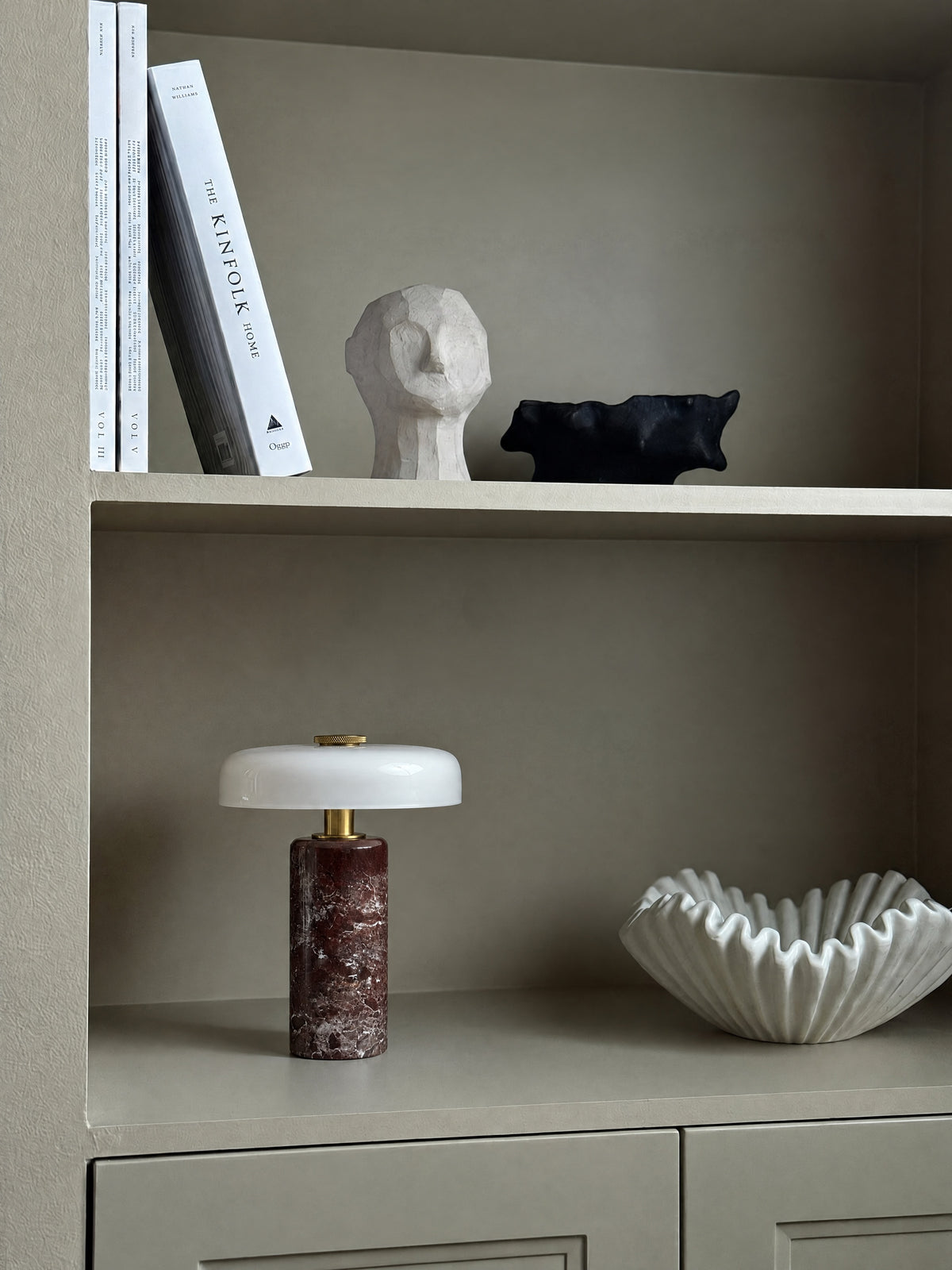 The Columba lamp unlit on a greige built-in shelf beside a white ruffled ceramic bowl, books and a carved head above.