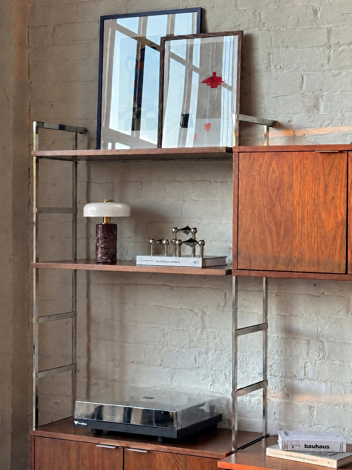The Columba lamp unlit on walnut and chrome wall shelving against a white brick wall, beside chrome candle holders.
