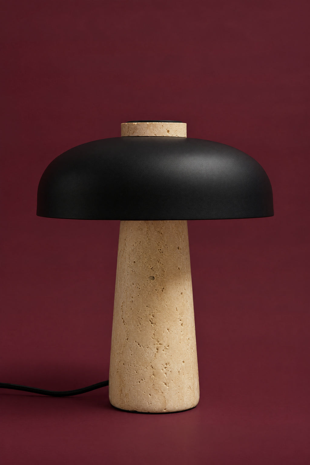 The Corvus lamp, unlit: a matte black metal dome on a warm honey travertine cone, its cord on the surface, against a deep oxblood background. (34 cm size.)