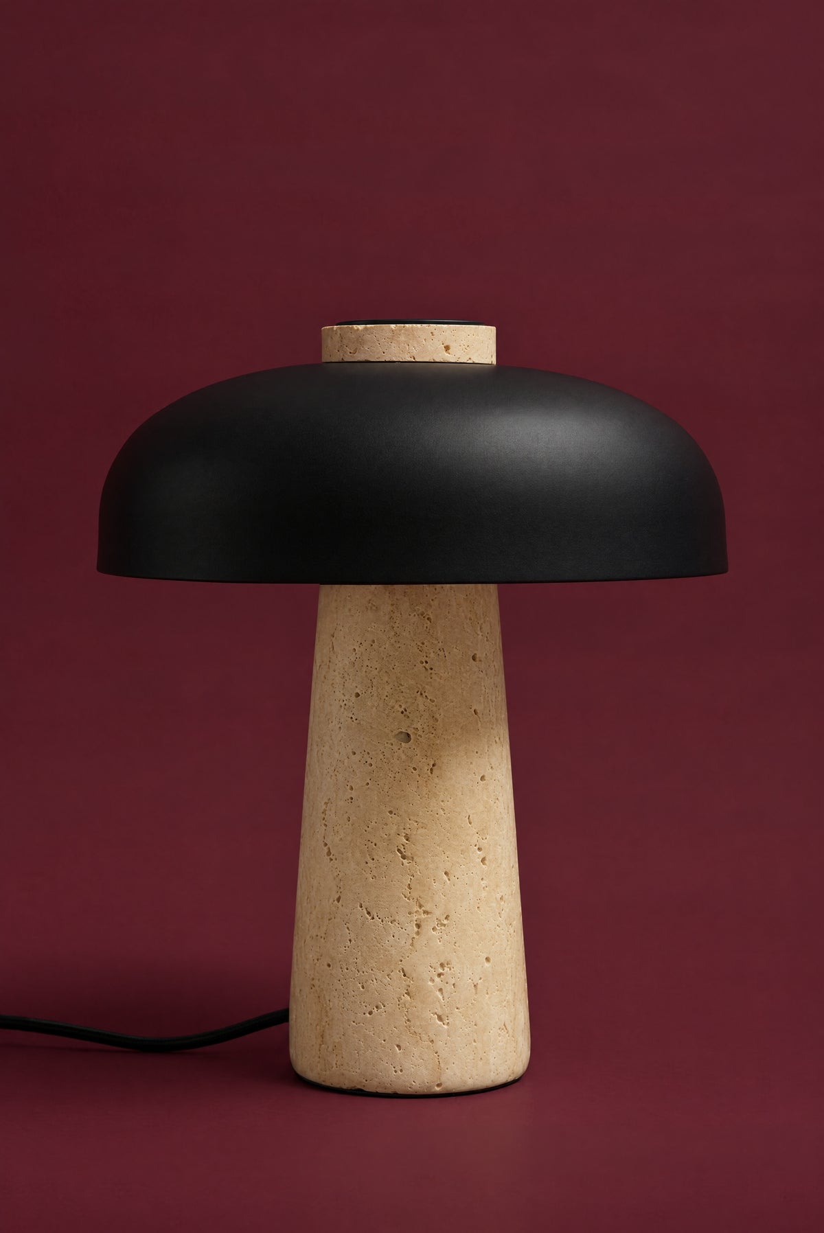 The Corvus lamp, unlit: a matte black metal dome on a warm honey travertine cone, its cord on the surface, against a deep oxblood background. (34 cm size.)