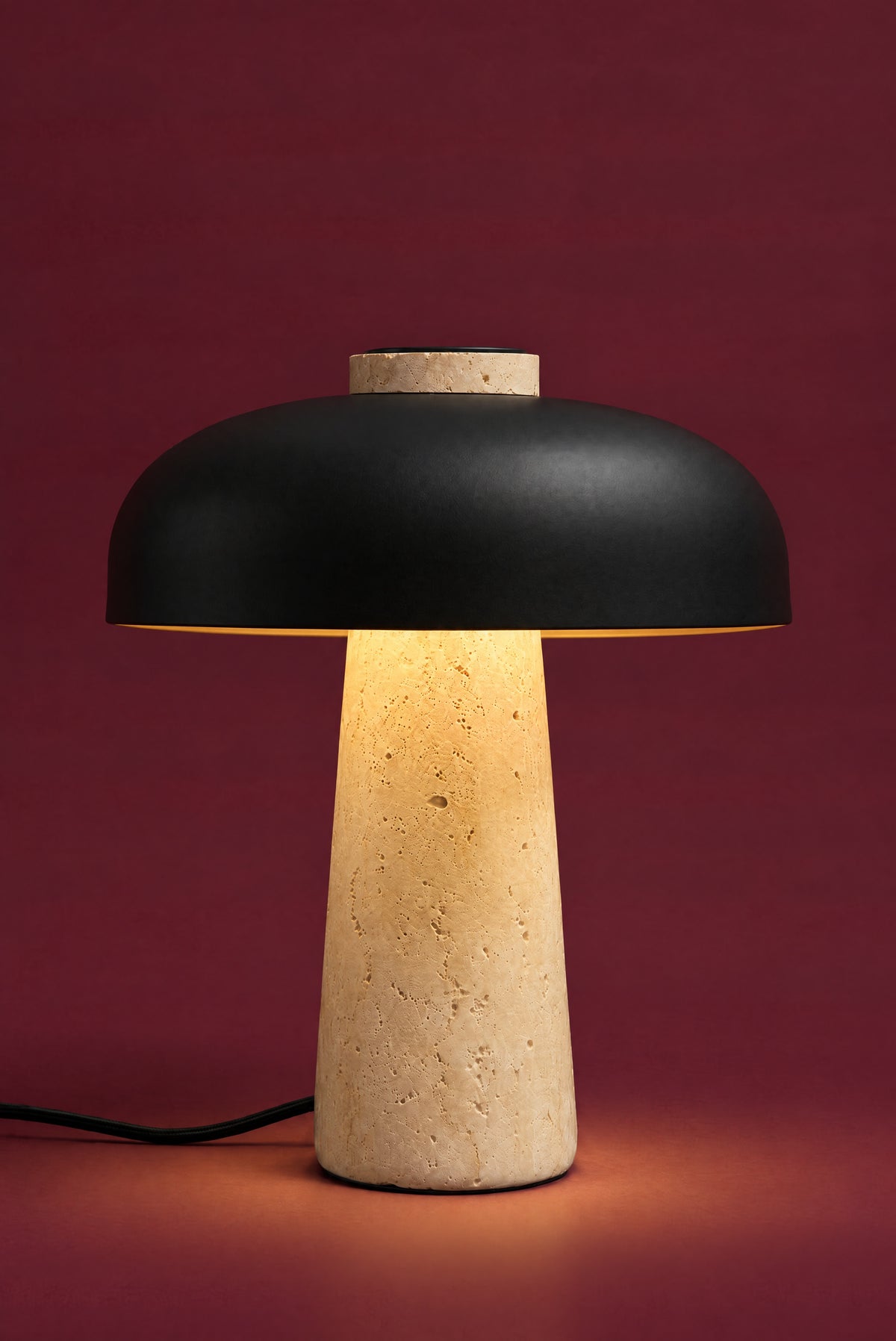 The Corvus lamp, lit: warm light glowing under the black dome and down the travertine cone, pooling on the surface. (34 cm size.)