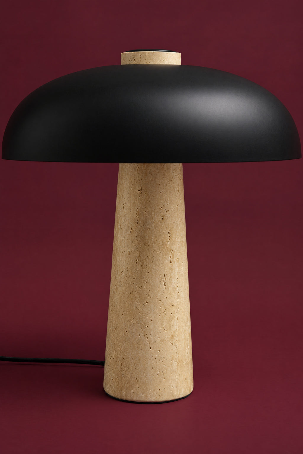 The Corvus lamp, unlit: a matte black metal dome on a tall warm honey travertine cone, its cord on the surface, against a deep oxblood background. (42 cm size.)