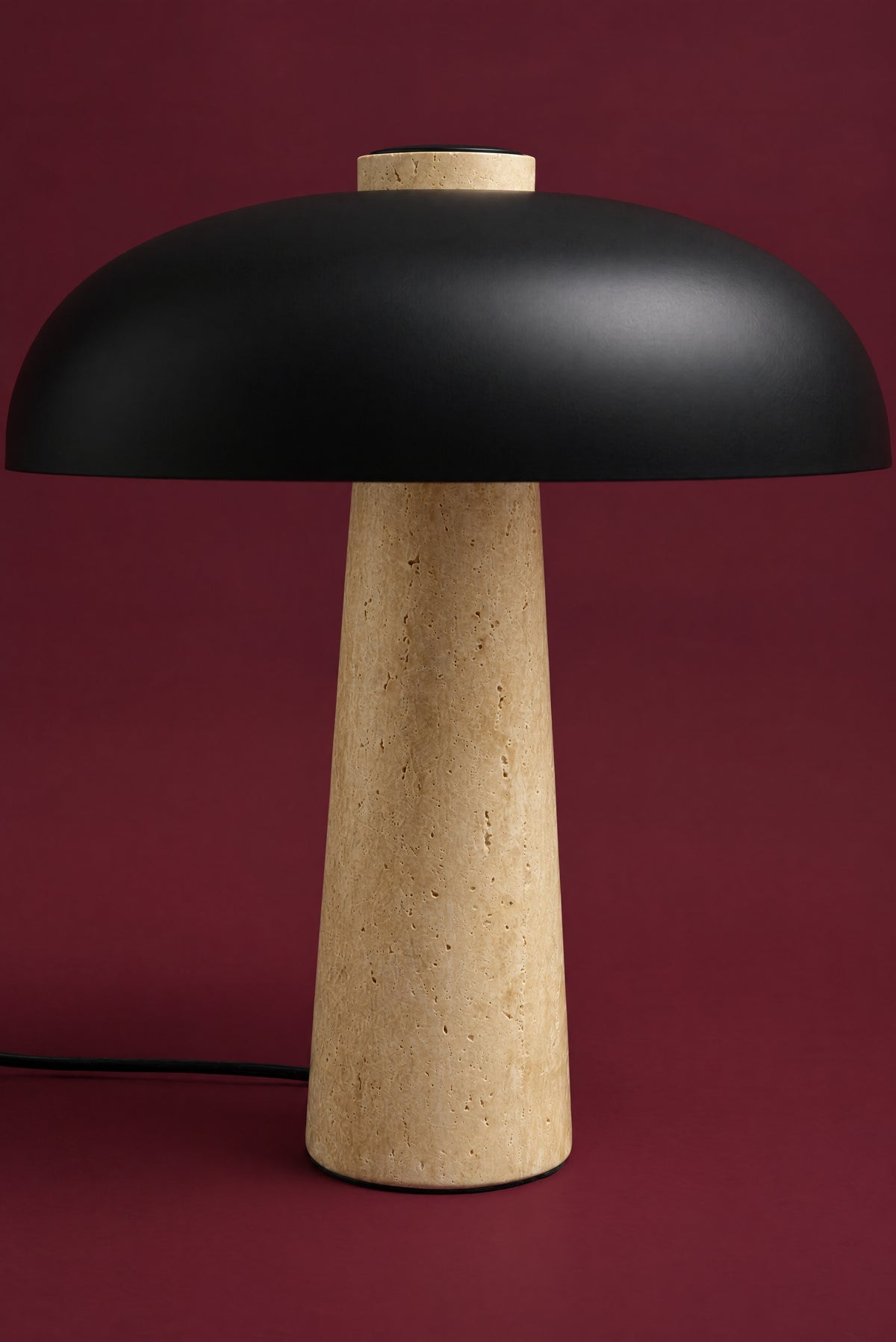 The Corvus lamp, unlit: a matte black metal dome on a tall warm honey travertine cone, its cord on the surface, against a deep oxblood background. (42 cm size.)