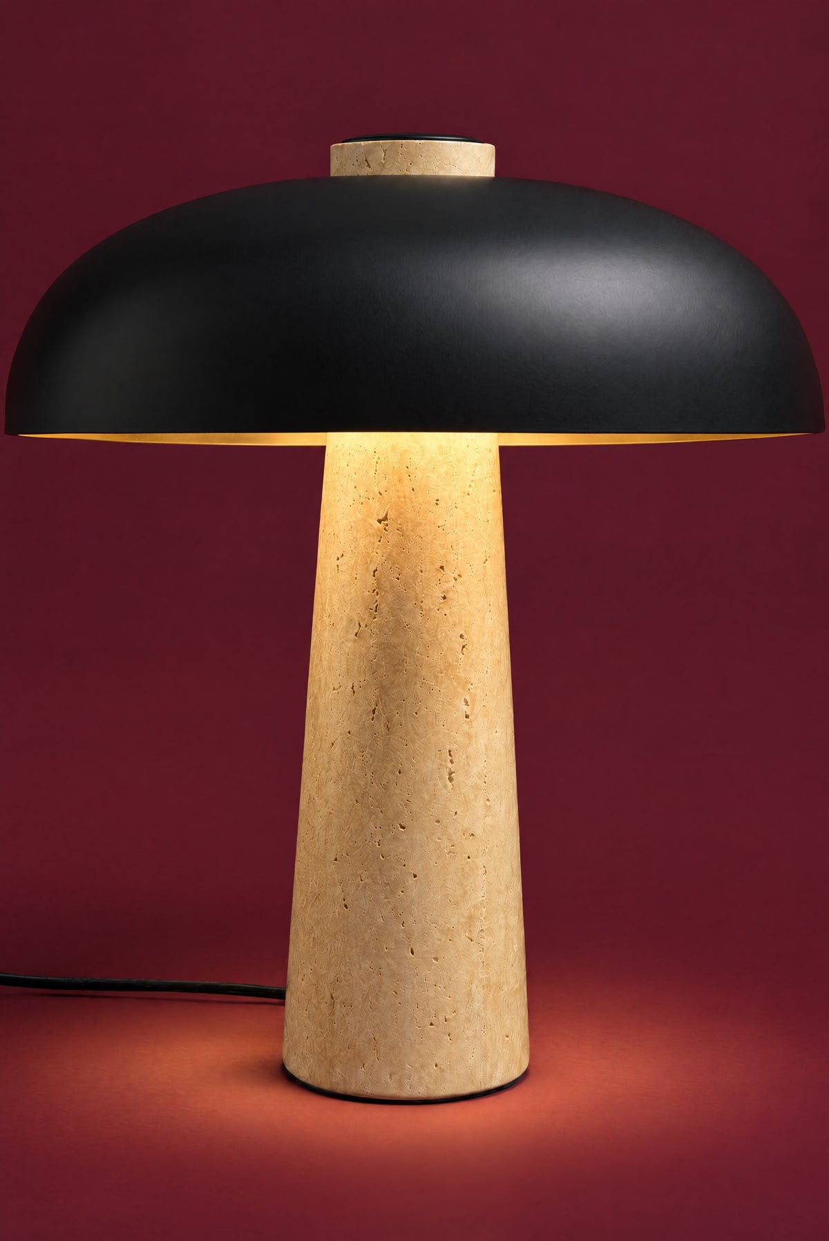 The Corvus lamp, lit: warm light glowing under the black dome and down the tall travertine cone, pooling on the surface. (42 cm size.)
