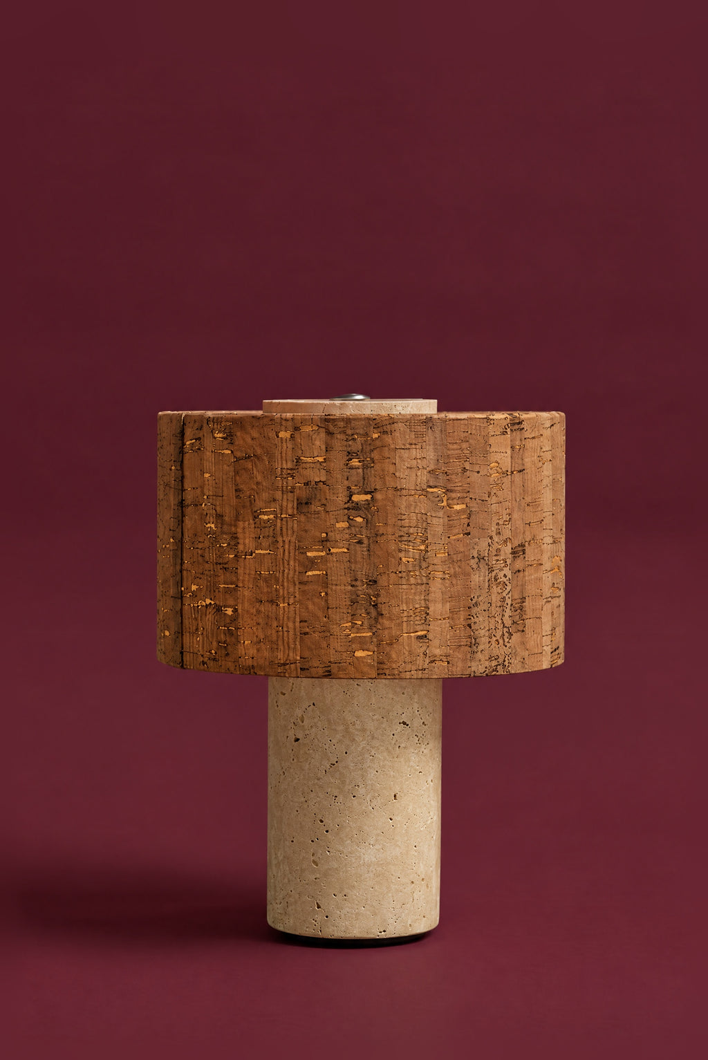 The Dorado lamp, unlit: a cork drum shade on a warm travertine column, against a deep oxblood background.