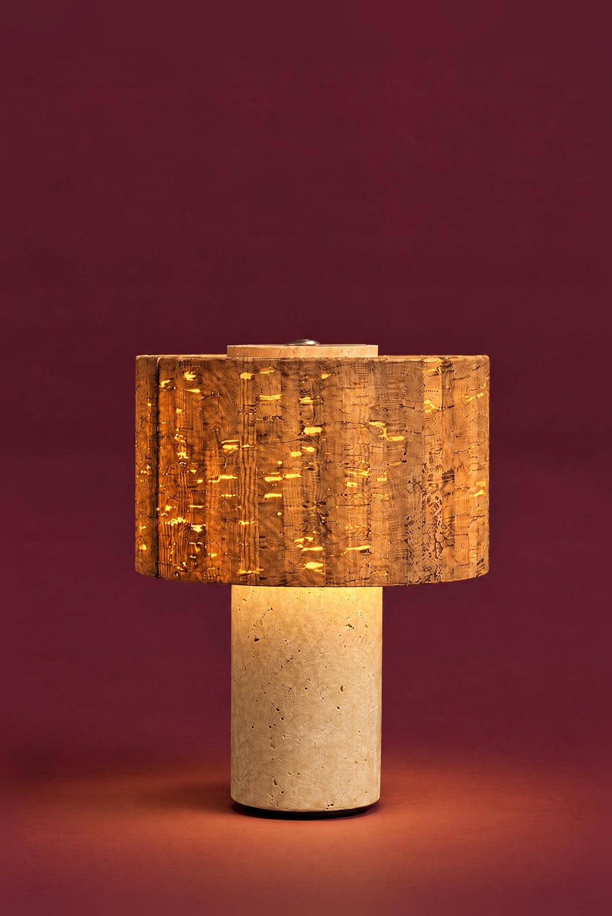 The Dorado lamp, lit: warm light glowing through the grain of the cork drum, a soft pool of light on the floor around the travertine column.