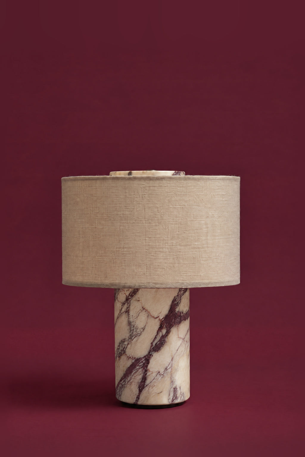 The Dorado lamp in linen, unlit: an oatmeal linen drum shade on a Calacatta Viola marble column with plum veining, against a deep oxblood background.