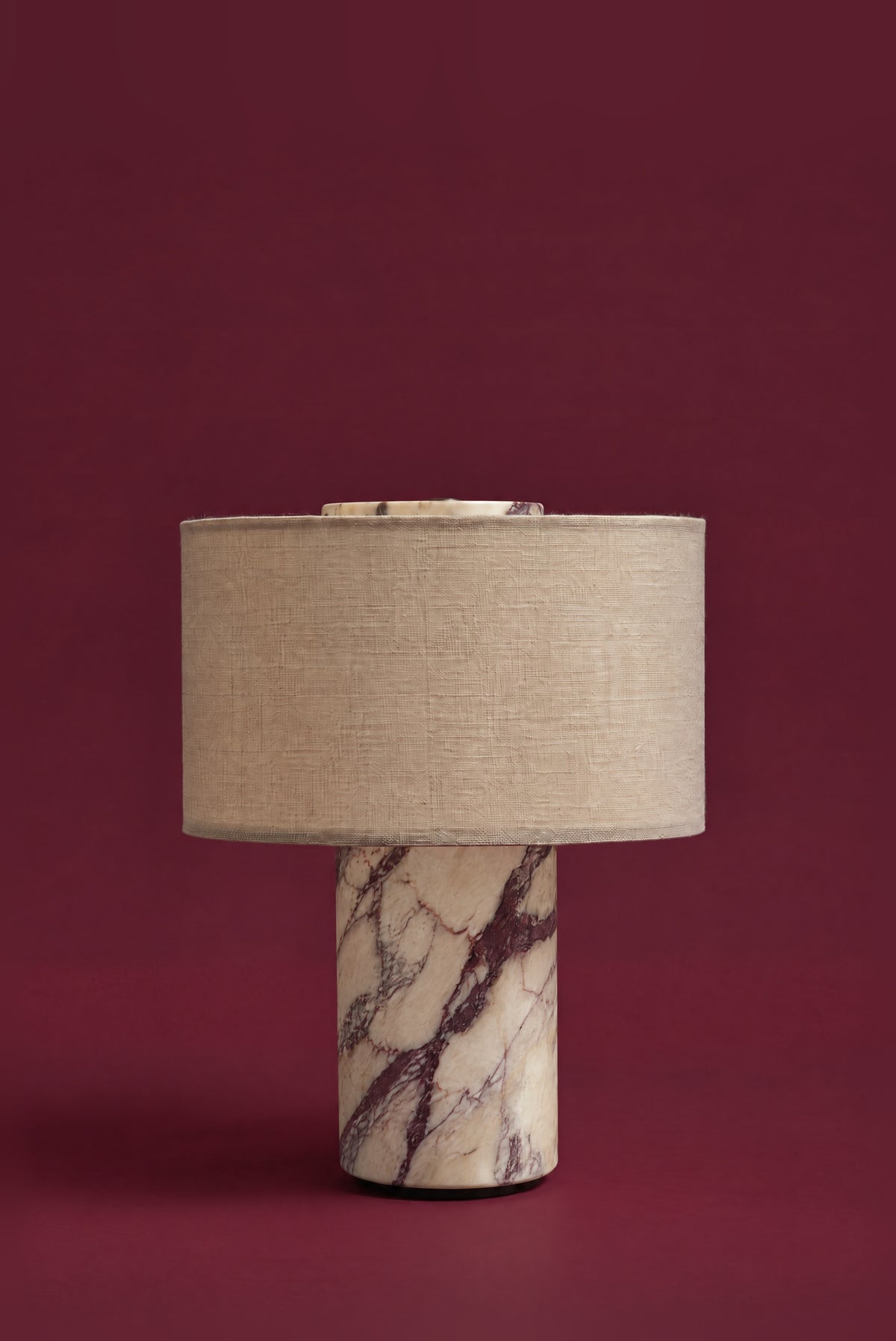 The Dorado lamp in linen, unlit: an oatmeal linen drum shade on a Calacatta Viola marble column with plum veining, against a deep oxblood background.