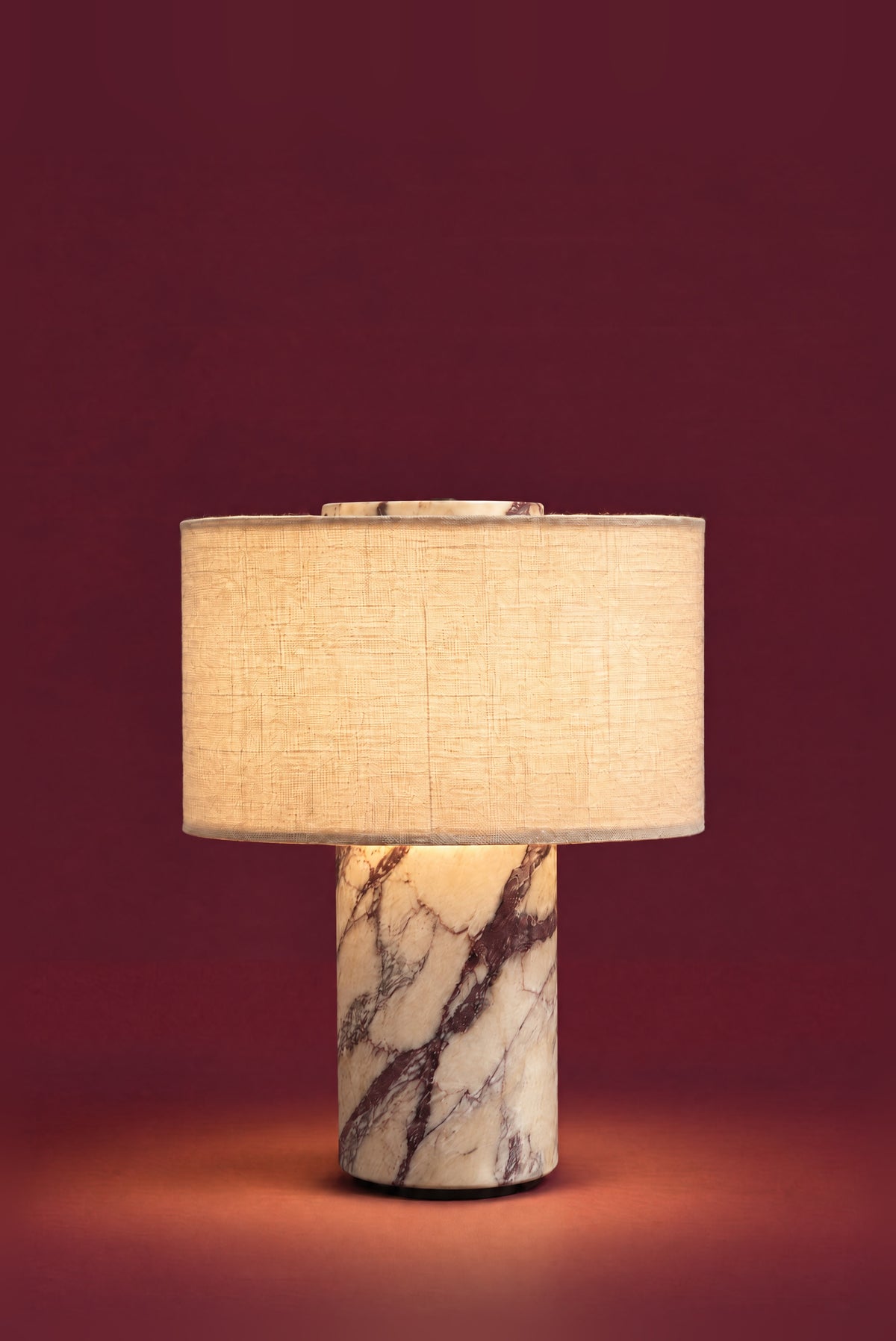 The Dorado lamp in linen, lit: the linen drum glowing warm and even, a soft pool of light on the floor around the veined marble column.