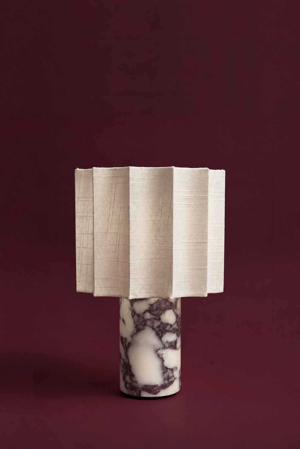 The Pavo lamp, unlit: a fluted white linen shade over a Calacatta Viola marble column with deep plum veins, against a deep oxblood background.