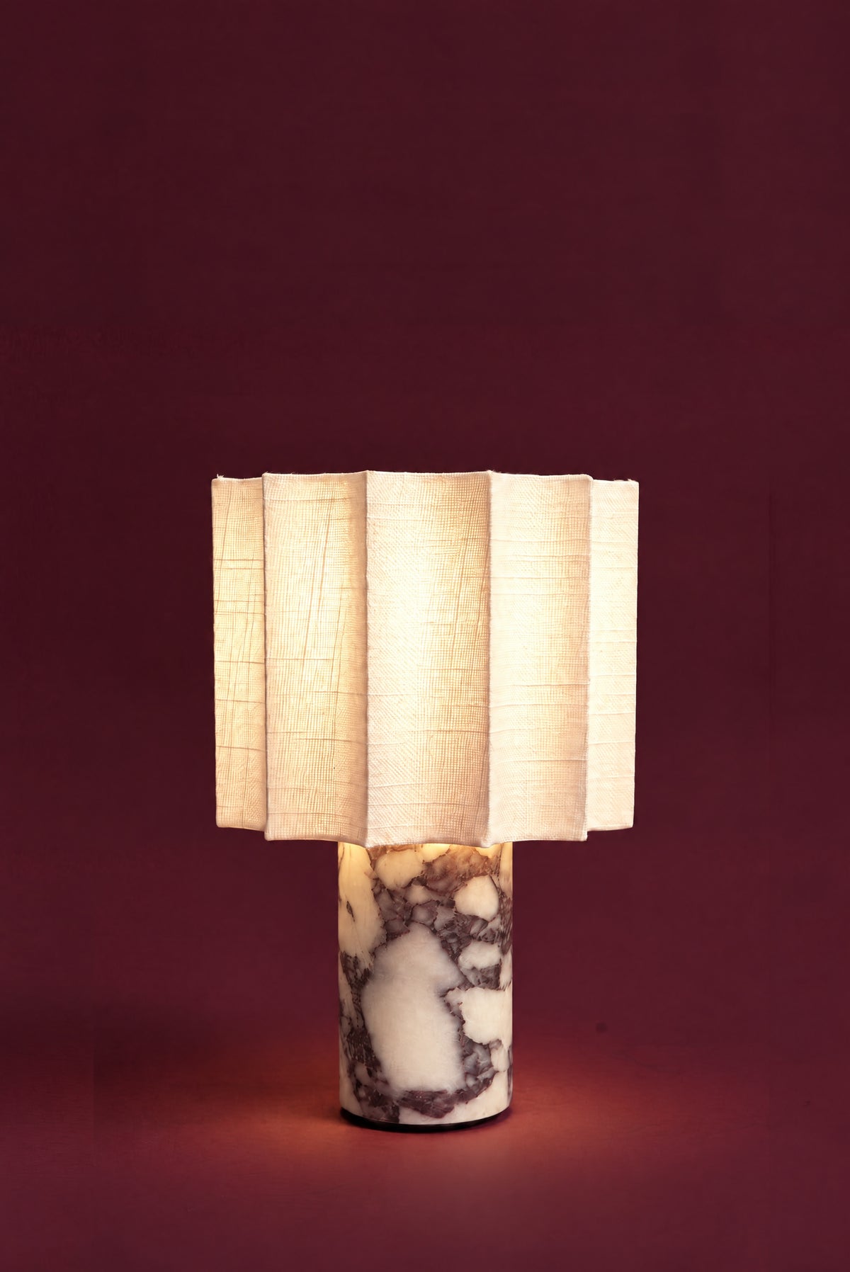 The Pavo lamp, lit: the fluted linen shade glowing warm white, light spilling onto the veined marble column and the floor.