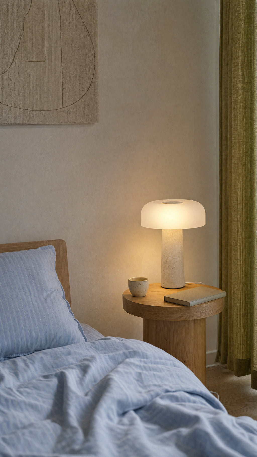 The Caelum lamp lit on an oak bedside table beside a striped blue pillow, a cup and a notebook, with a green linen curtain.