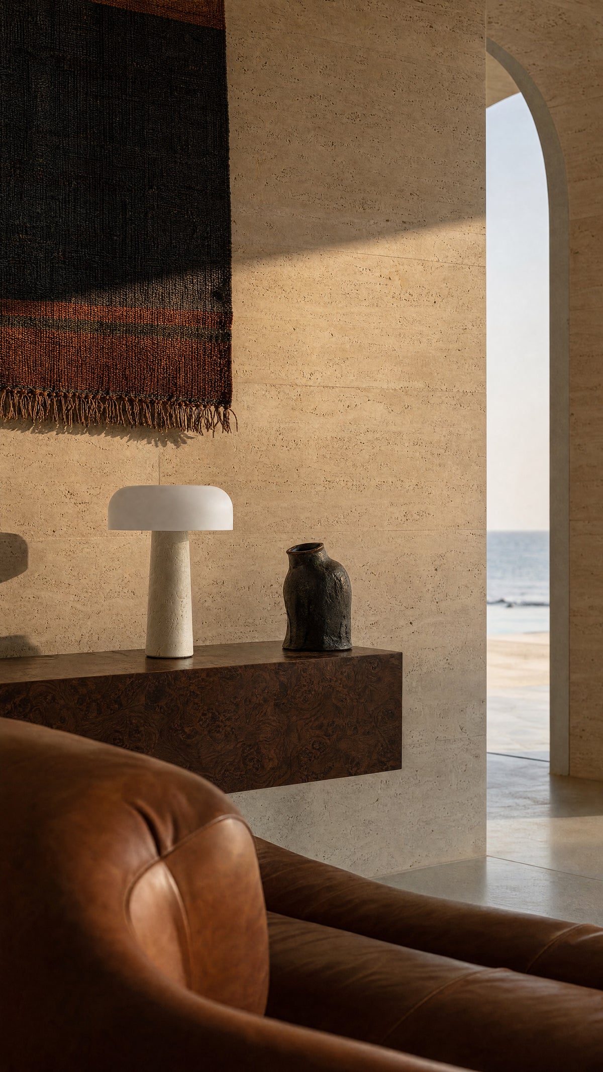 The Caelum lamp unlit on a burl-wood console against a travertine wall, a leather sofa in front and an arch opening to the sea.