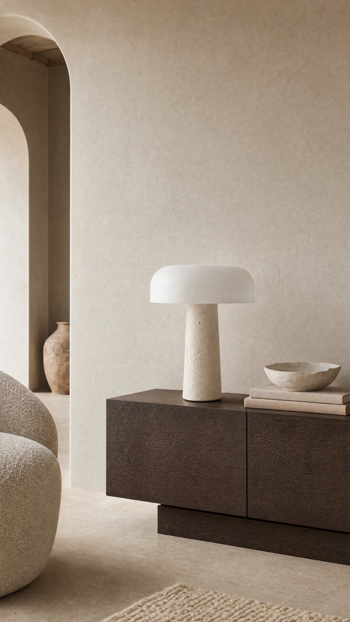 The Caelum lamp unlit on a dark oak sideboard in a plaster-walled living room, a stone bowl on a stack of books beside it.