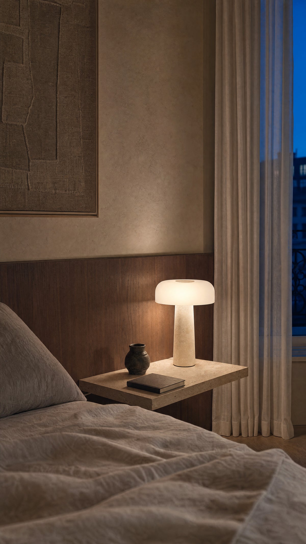 The Caelum lamp lit on a travertine bedside shelf at dusk, wood panelling behind and sheer curtains at the window.