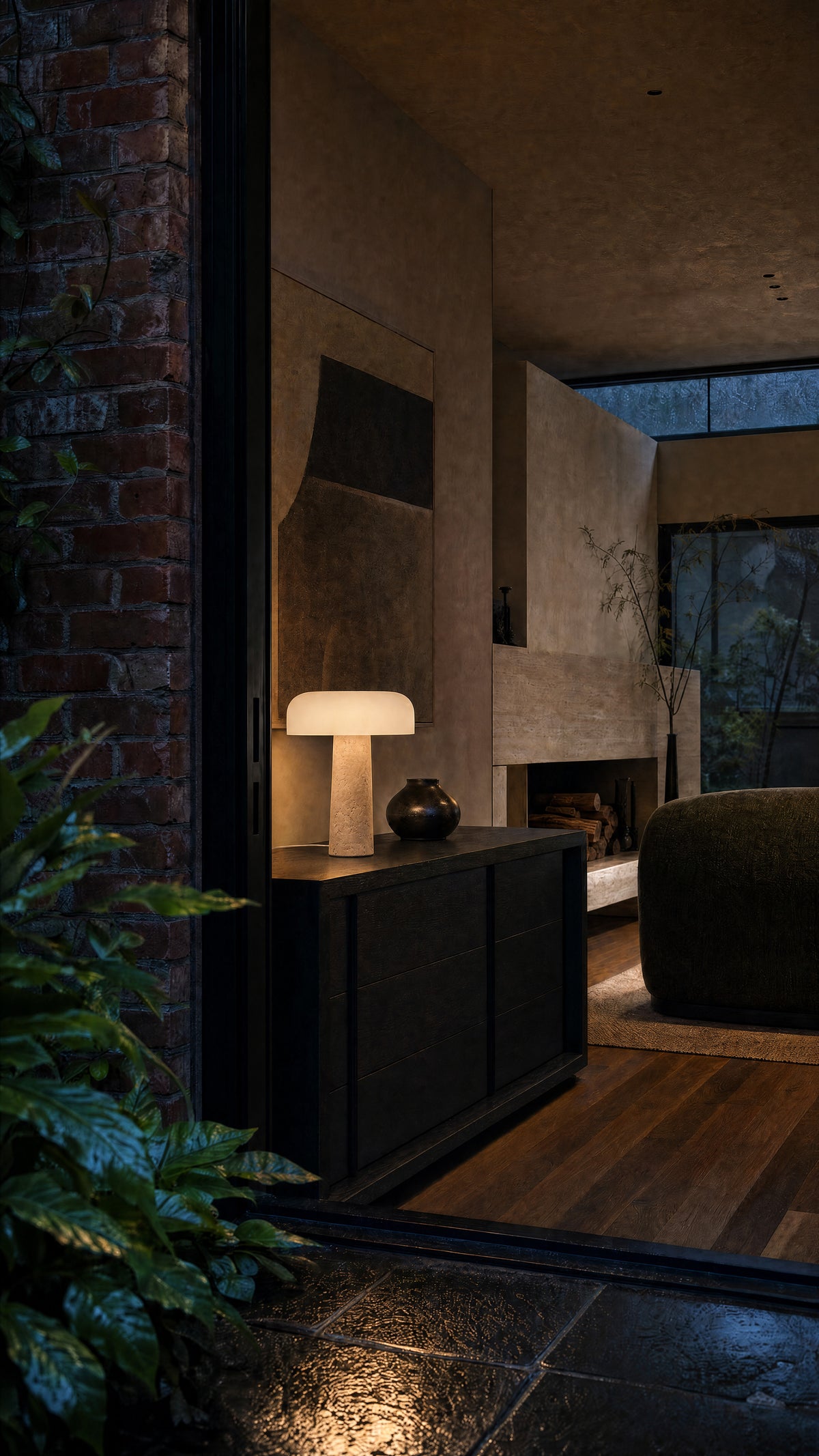 The Caelum lamp lit on a black oak sideboard seen through a doorway at blue hour, a brick wall and wet leaves in the foreground, a fireplace beyond.