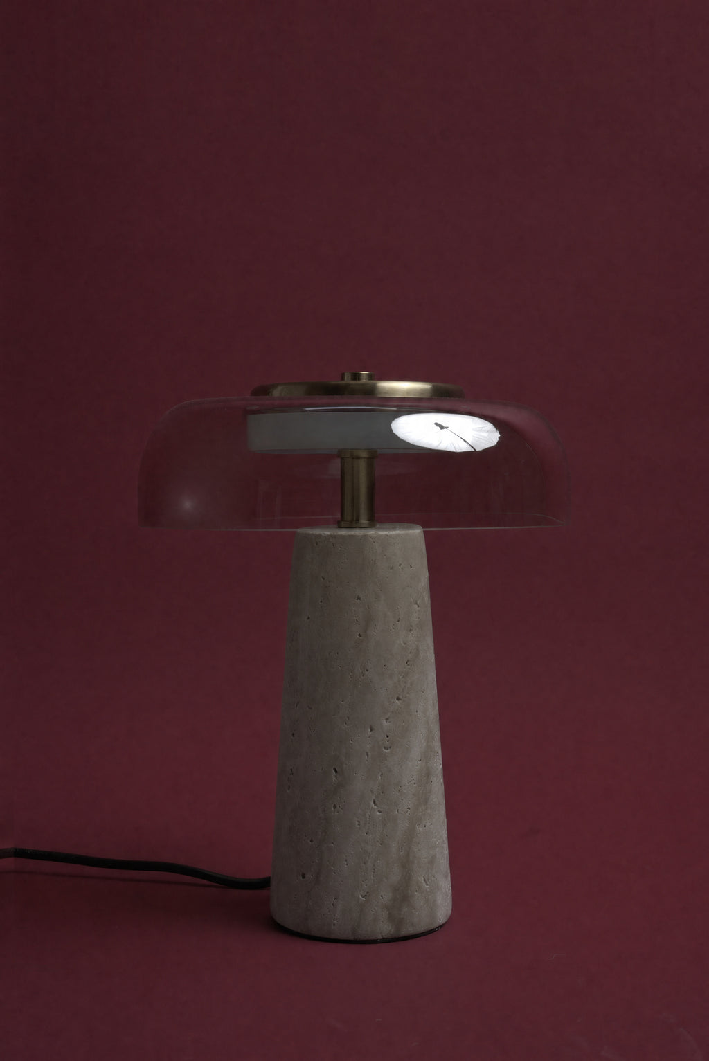 The Ankaa lamp, unlit: a transparent smoked rose glass dome on a pale travertine cone, cord visible, against a deep oxblood background.
