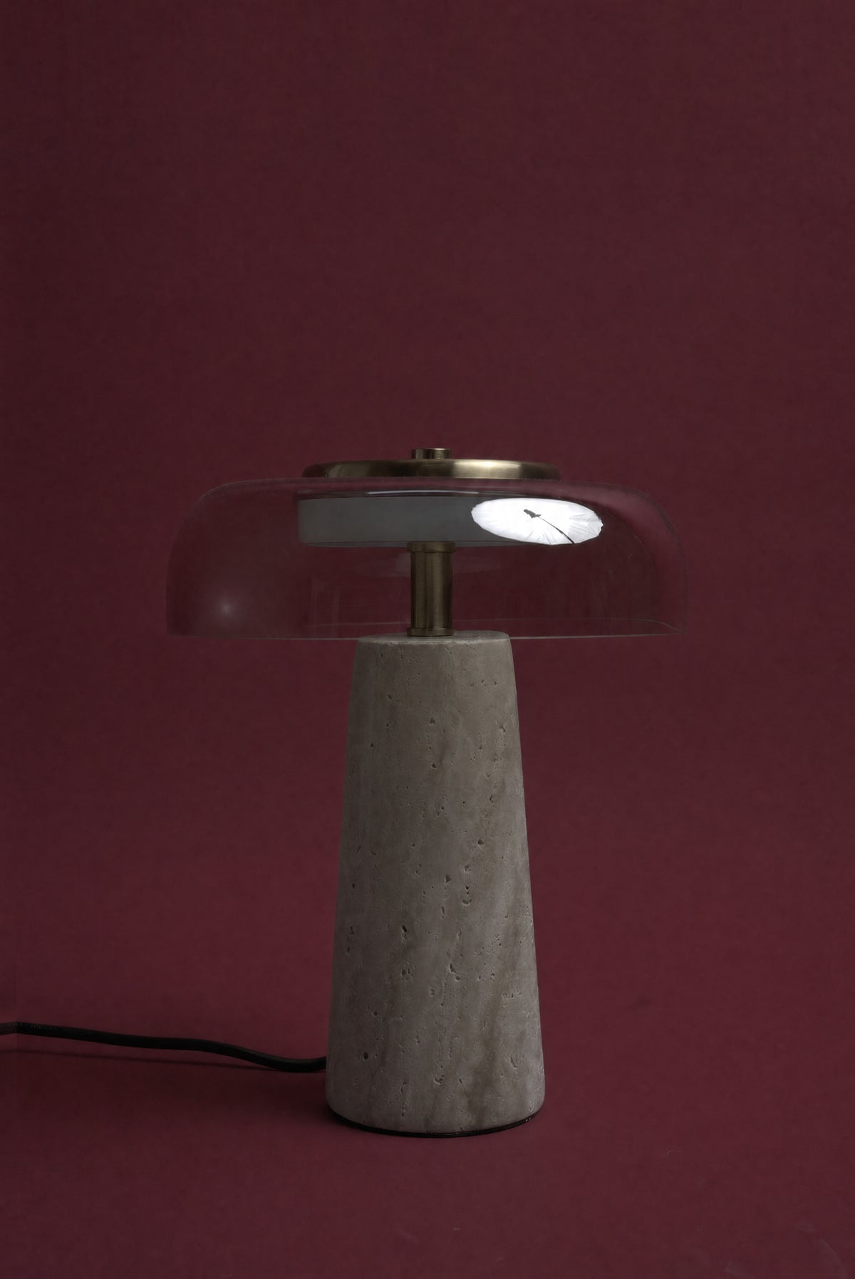 The Ankaa lamp, unlit: a transparent smoked rose glass dome on a pale travertine cone, cord visible, against a deep oxblood background.