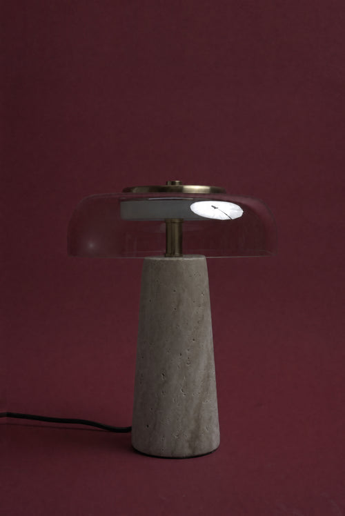 The Ankaa lamp, unlit: a transparent smoked rose glass dome on a pale travertine cone, cord visible, against a deep oxblood background.