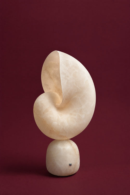 The Volans lamp, unlit: a nautilus shell carved from warm cream alabaster on a rounded alabaster foot with a small chrome touch button, against a deep oxblood background.